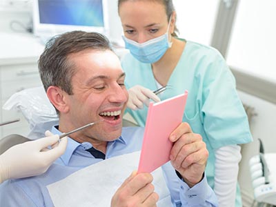 The image shows a man in a dental office, smiling and holding up a pink card with the word Congratulations written on it. He is seated in a chair with medical equipment around him, wearing a surgical mask, and there s a female dental professional in the background assisting him.